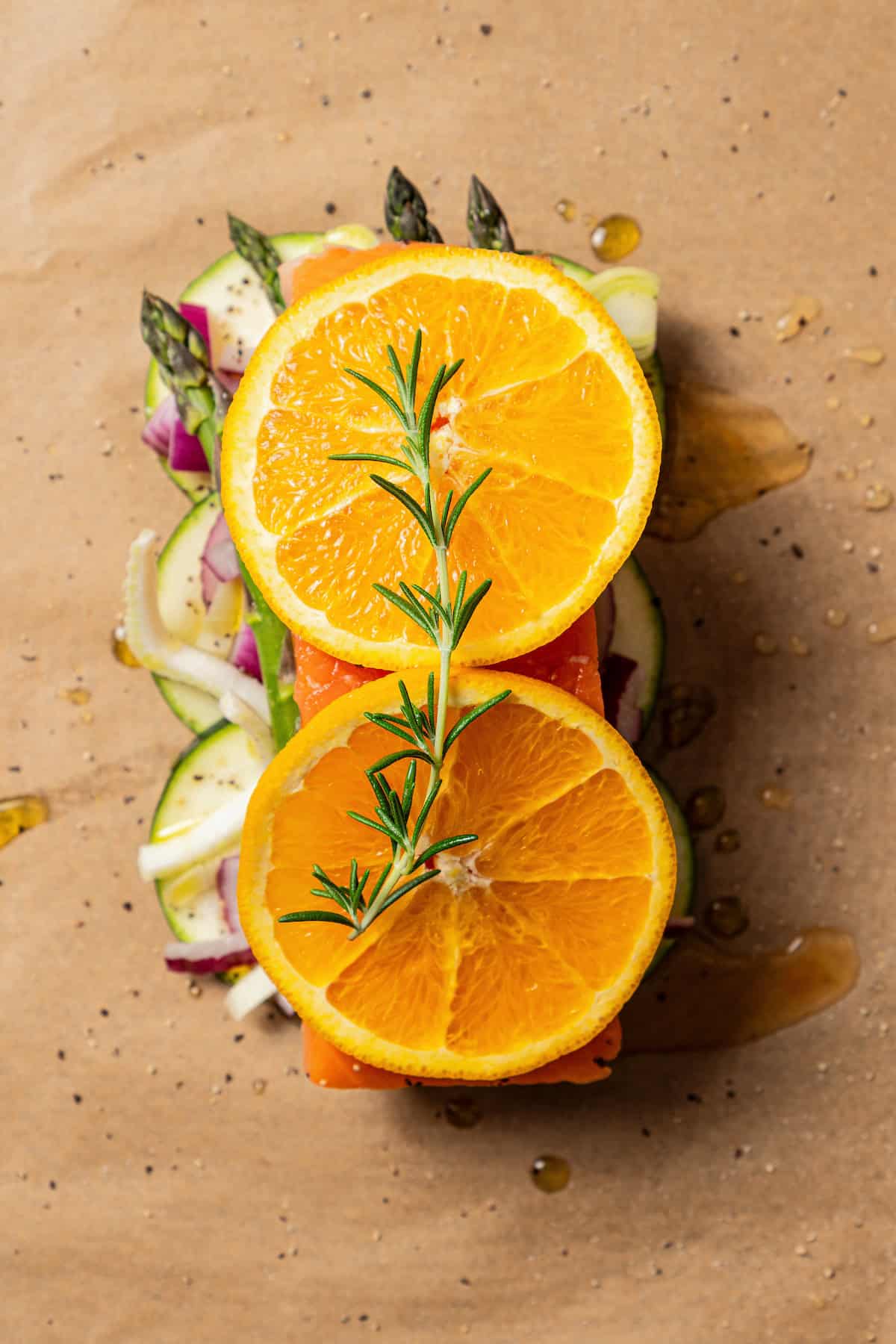 Overhead view of a salmon filet nestled over top of chopped vegetables on parchment paper, topped with orange slices and a thyme sprig.