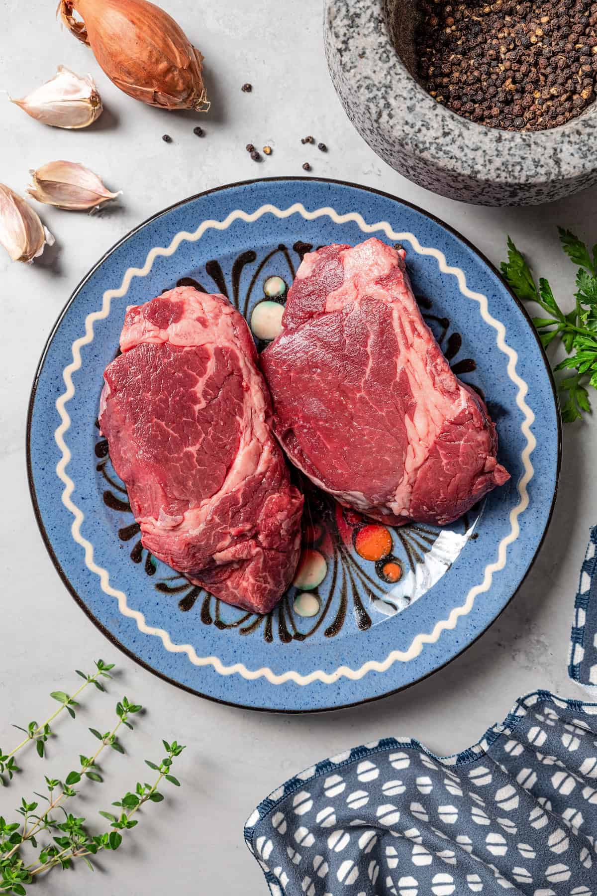 Overhead view of two steaks on a blue patterned plate next to a mortar filled with crushed peppercorns.