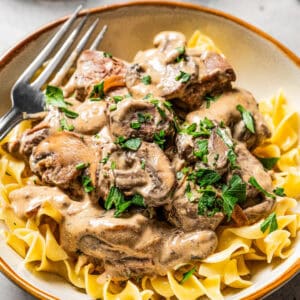 A bowl of beef stroganoff garnished with parsley with a fork, and a bowl of mushrooms in the background.