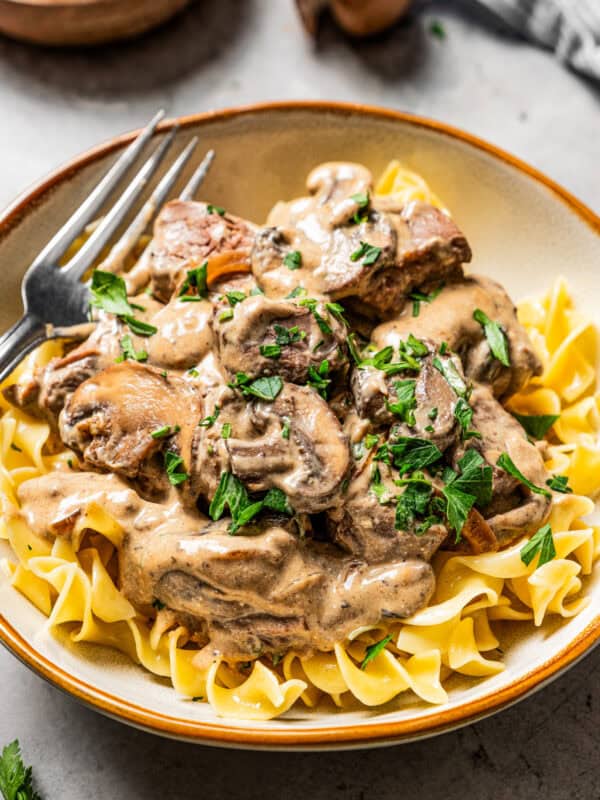 A bowl of beef stroganoff garnished with parsley with a fork, and a bowl of mushrooms in the background.