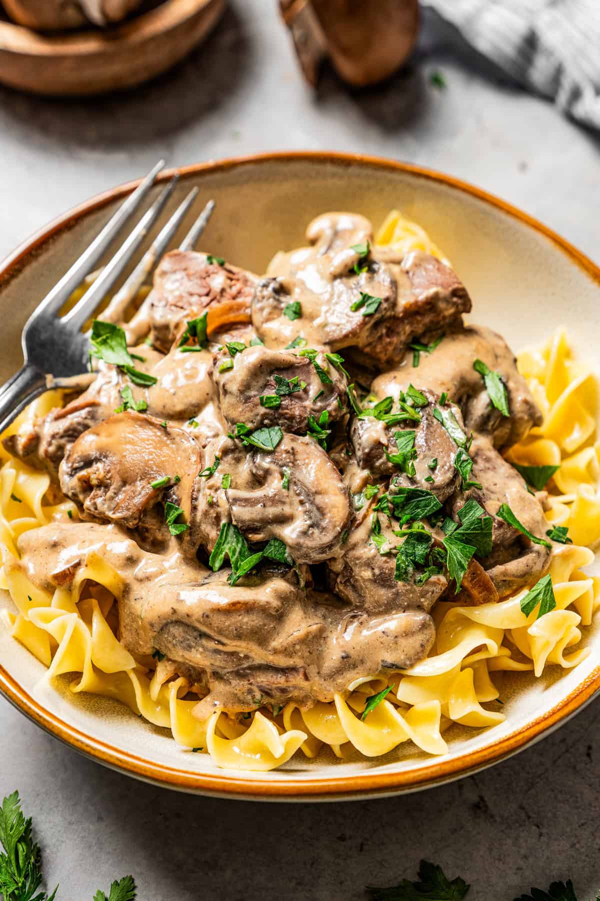 A bowl of beef stroganoff garnished with parsley with a fork, and a bowl of mushrooms in the background.