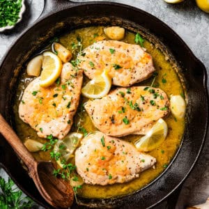 Overhead view of chicken paillard nestled into a large skillet with sauce, garnished with lemon wedges and fresh parsley.