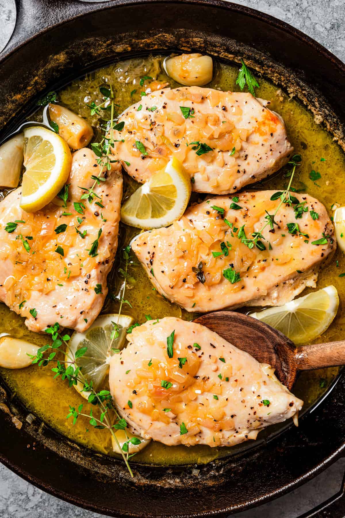 Four cooked chicken breasts in a skillet, garnished with lemon slices and chopped parsley.