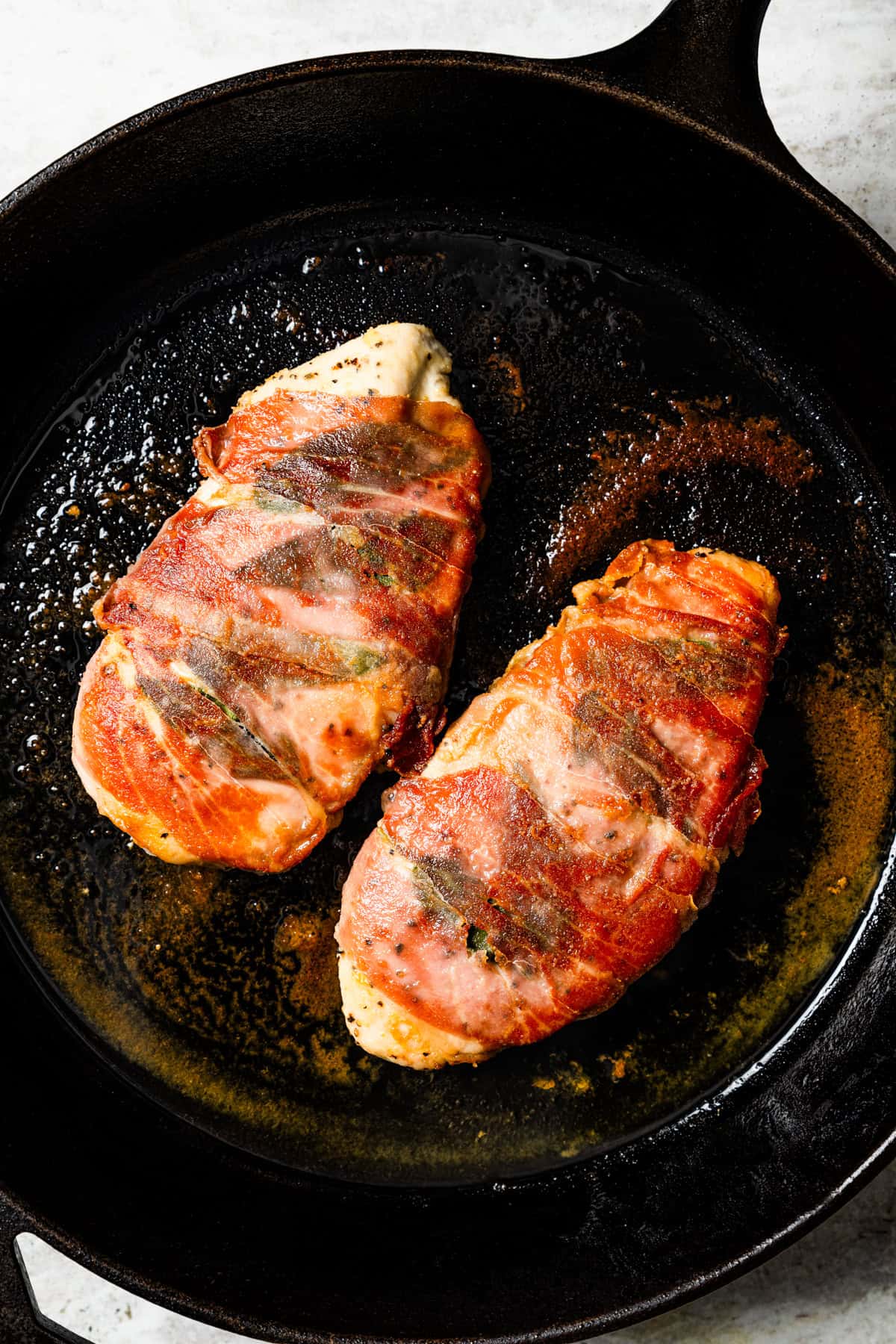 Two chicken breasts wrapped in prosciutto in a cast iron skillet.