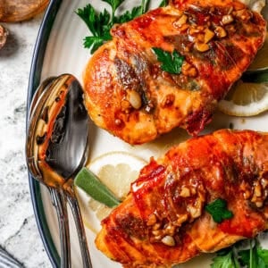 Two pieces of chicken saltimbocca garnished with parsley and lemon slices are served on a platter next to a spoon and a jar of sauce.