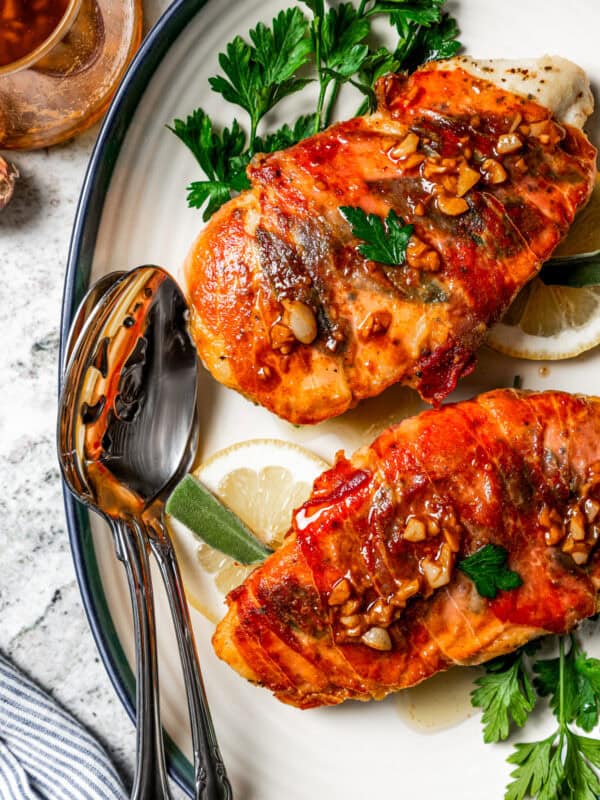 Two pieces of chicken saltimbocca garnished with parsley and lemon slices are served on a platter next to a spoon and a jar of sauce.