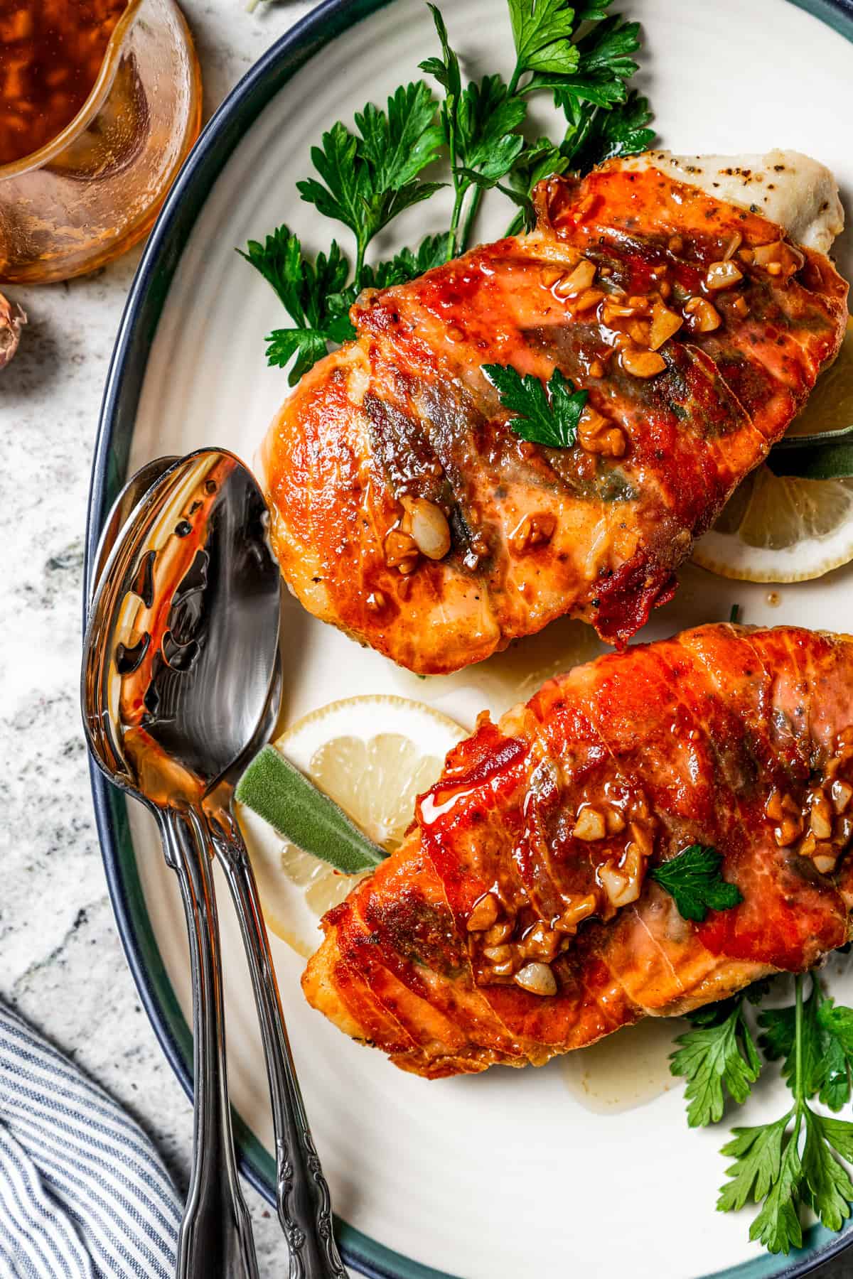 Two pieces of chicken saltimbocca garnished with parsley and lemon slices are served on a platter next to a spoon and a jar of sauce.