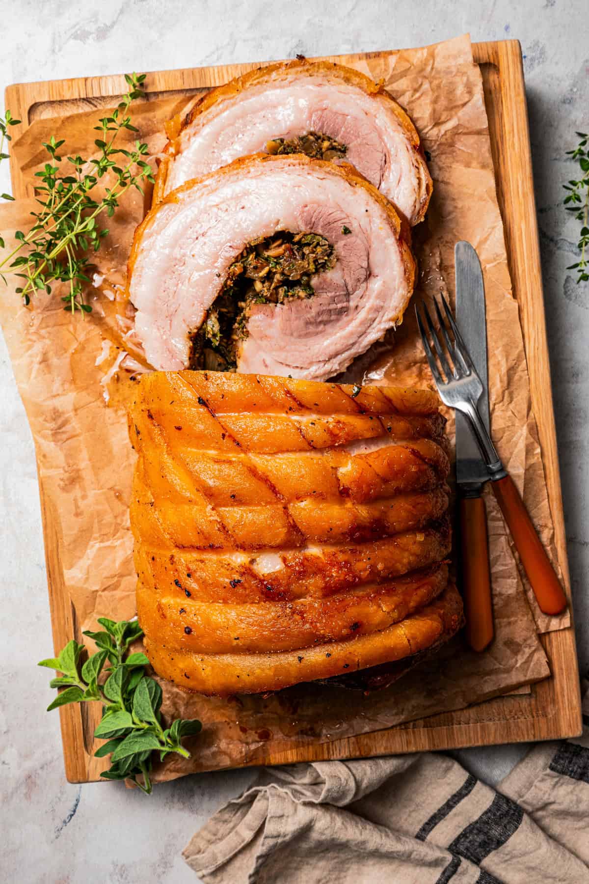 Overhead view of sliced porchetta on a wooden cutting board, next to fresh herb sprigs and a knife and fork.