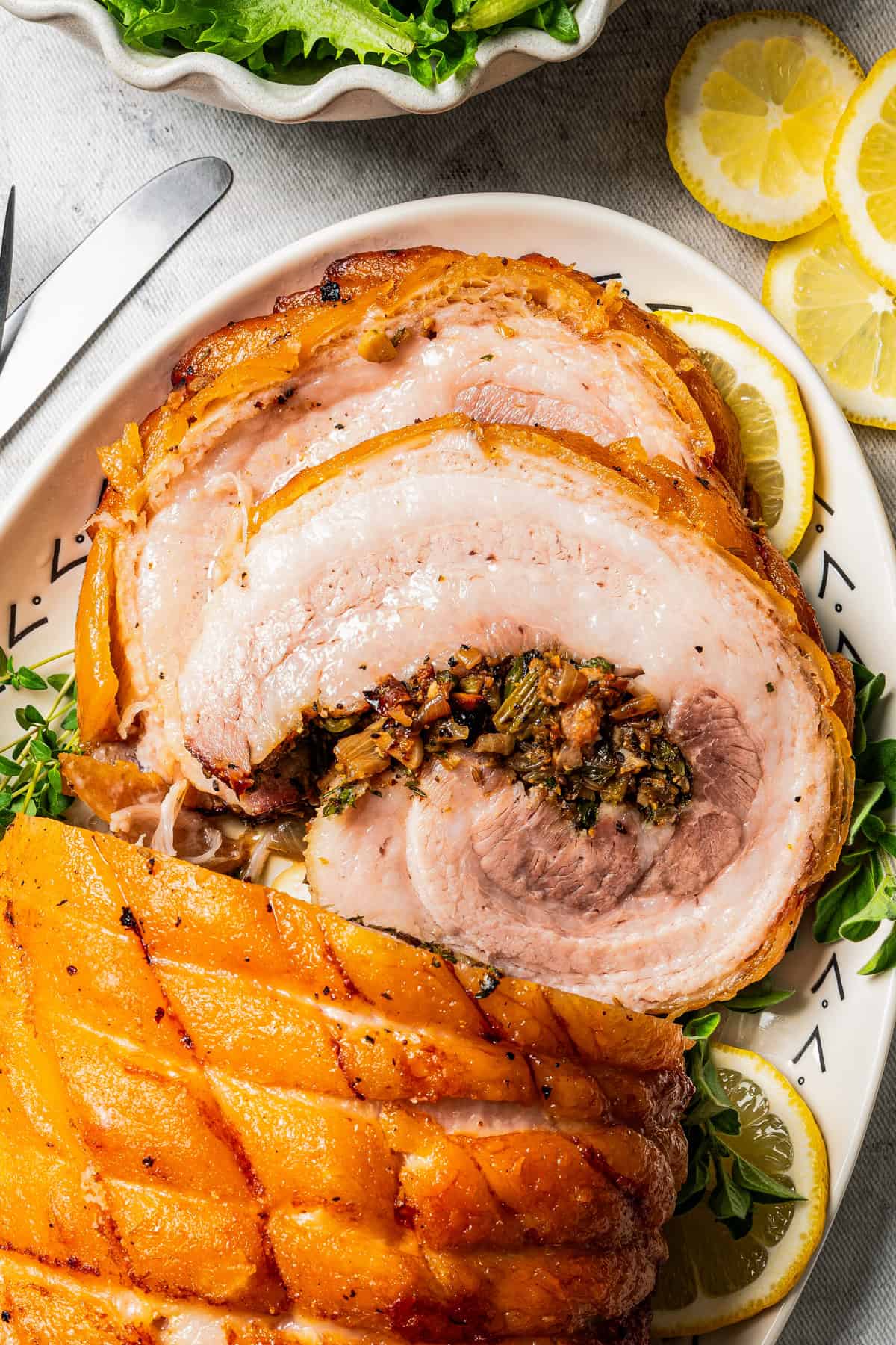 Overhead close up view of sliced porchetta on a platter next to a bowl of fresh greens.