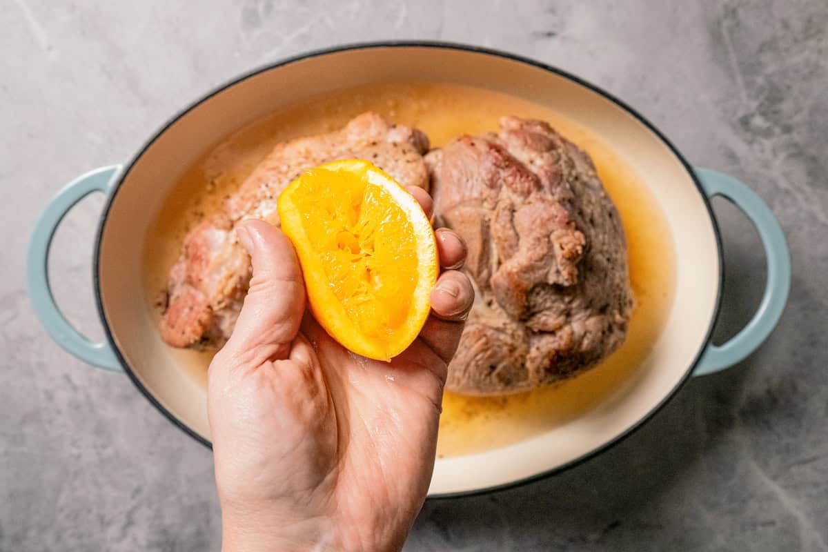 This pork roast recipe is a whole meal in one! With juicy seasoned pork shoulder slow-roasted with fresh citrus, carrots, potatoes, and herbs. A hand squeezing an orange over a pork shoulder roast.