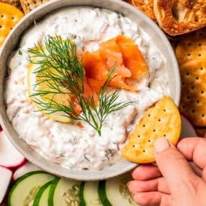 Hand dipping a cracker into a bowl of dip garnished with smoked salmon, lemon, and a fresh dill sprig, surrounded by a platter of crackers and crudités.