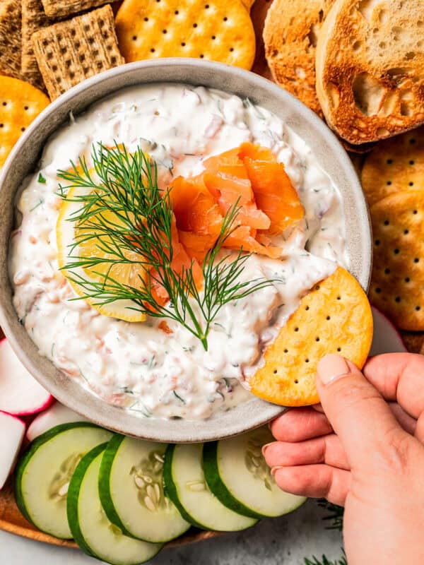 Hand dipping a cracker into a bowl of dip garnished with smoked salmon, lemon, and a fresh dill sprig, surrounded by a platter of crackers and crudités.