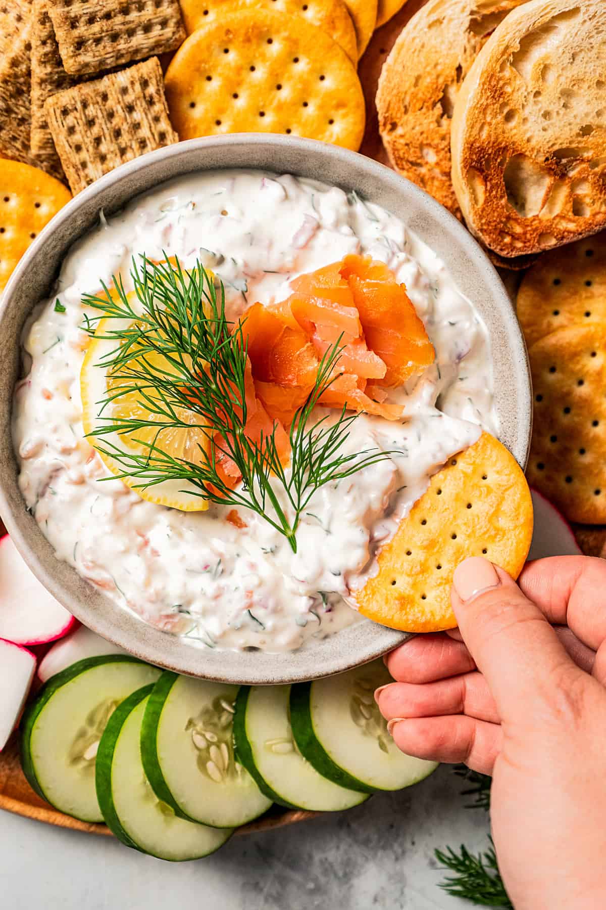 Hand dipping a cracker into a bowl of dip garnished with smoked salmon, lemon, and a fresh dill sprig, surrounded by a platter of crackers and crudités.