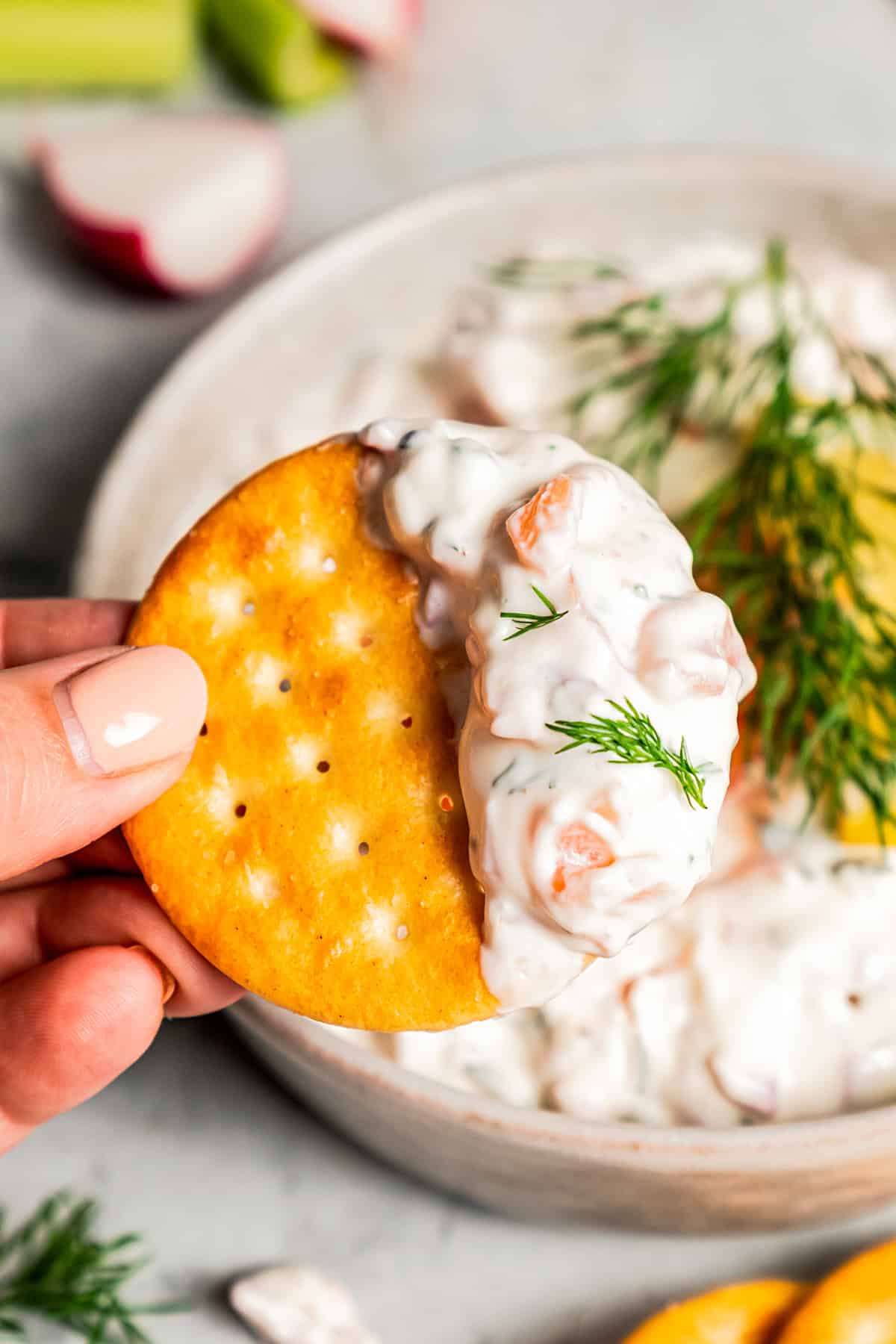 Close-up of a hand holding up a cracker dipped in smoked salmon dip with a bowl of dip in the background.