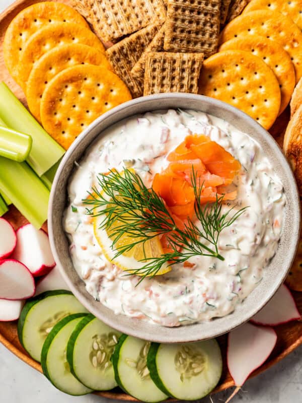 Overhead view of a bowl of smoked salmon dip garnished with lox, lemon, and a fresh dill sprig, surrounded by crackers and crudités on a platter.