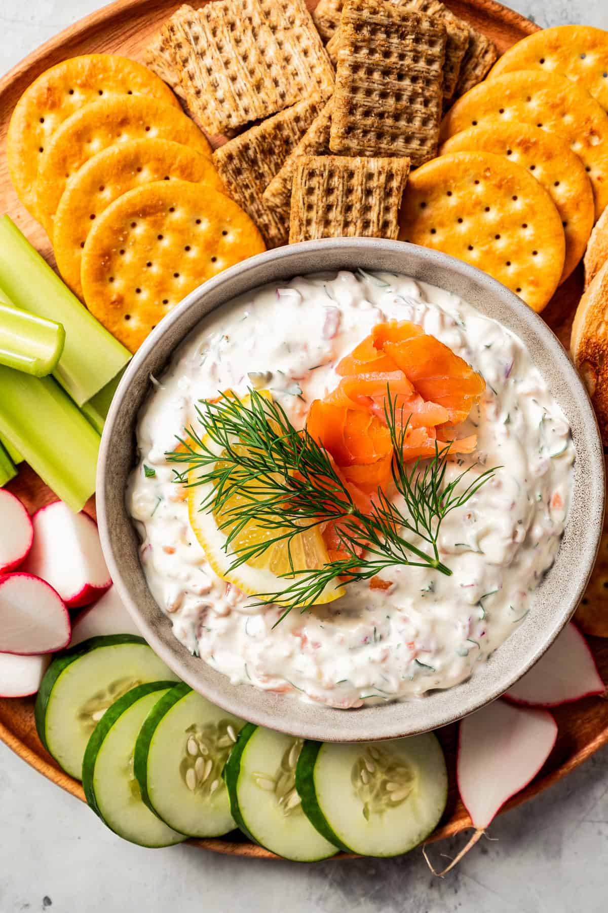 Overhead view of a bowl of smoked salmon dip garnished with lox, lemon, and a fresh dill sprig, surrounded by crackers and crudités on a platter.