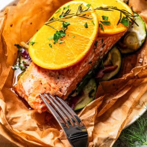 A fork pokes into the end of a salmon filet garnished with orange slices in parchment paper.