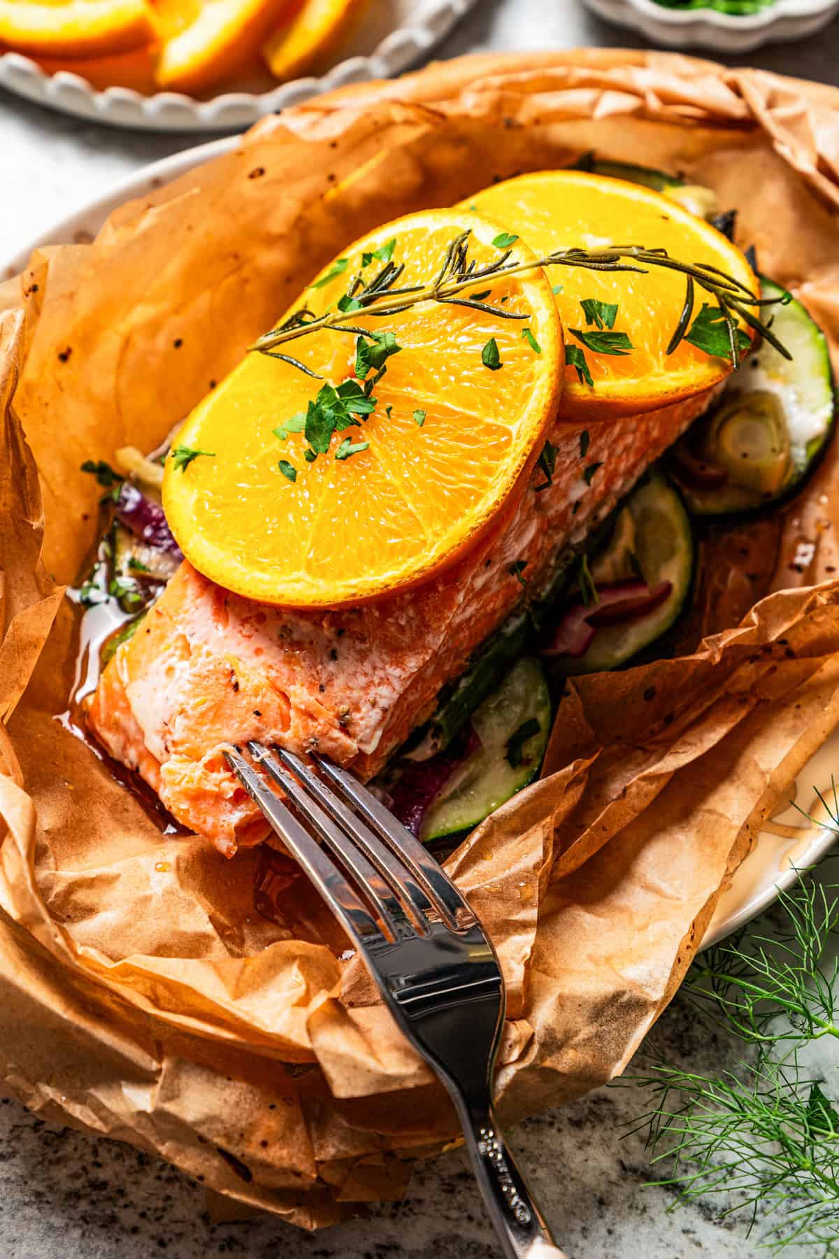 A fork pokes into the end of a salmon filet garnished with orange slices in parchment paper.