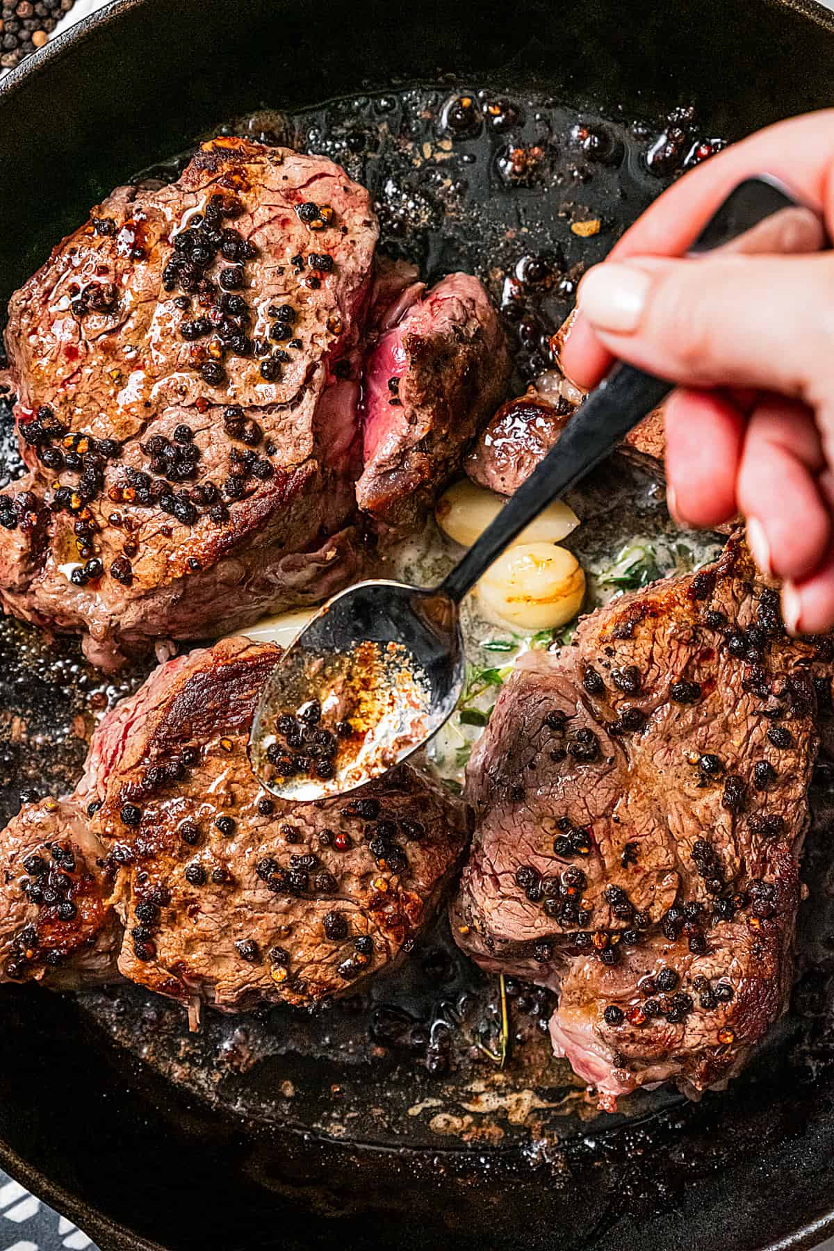 A hand uses a spoon to drizzle seasoned butter over peppercorn-crusted steaks as they cook in a cast iron skillet.