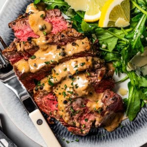 Sliced steak au poivre drizzled with cognac sauce is served on a plate next to a serving of green salad garnished with lemon wedges, and a fork is placed on the side.