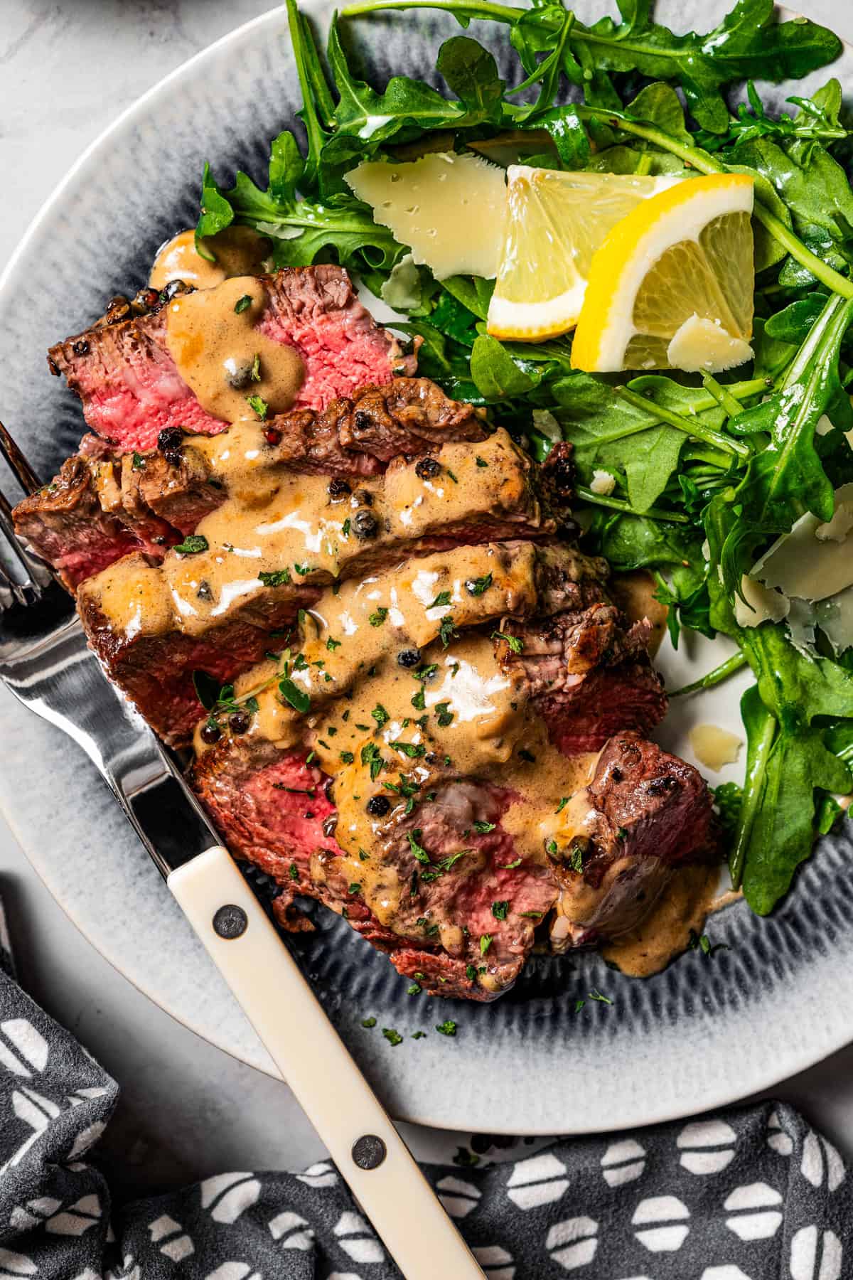 Sliced steak au poivre drizzled with cognac sauce is served on a plate next to a serving of green salad garnished with lemon wedges, and a fork is placed on the side.