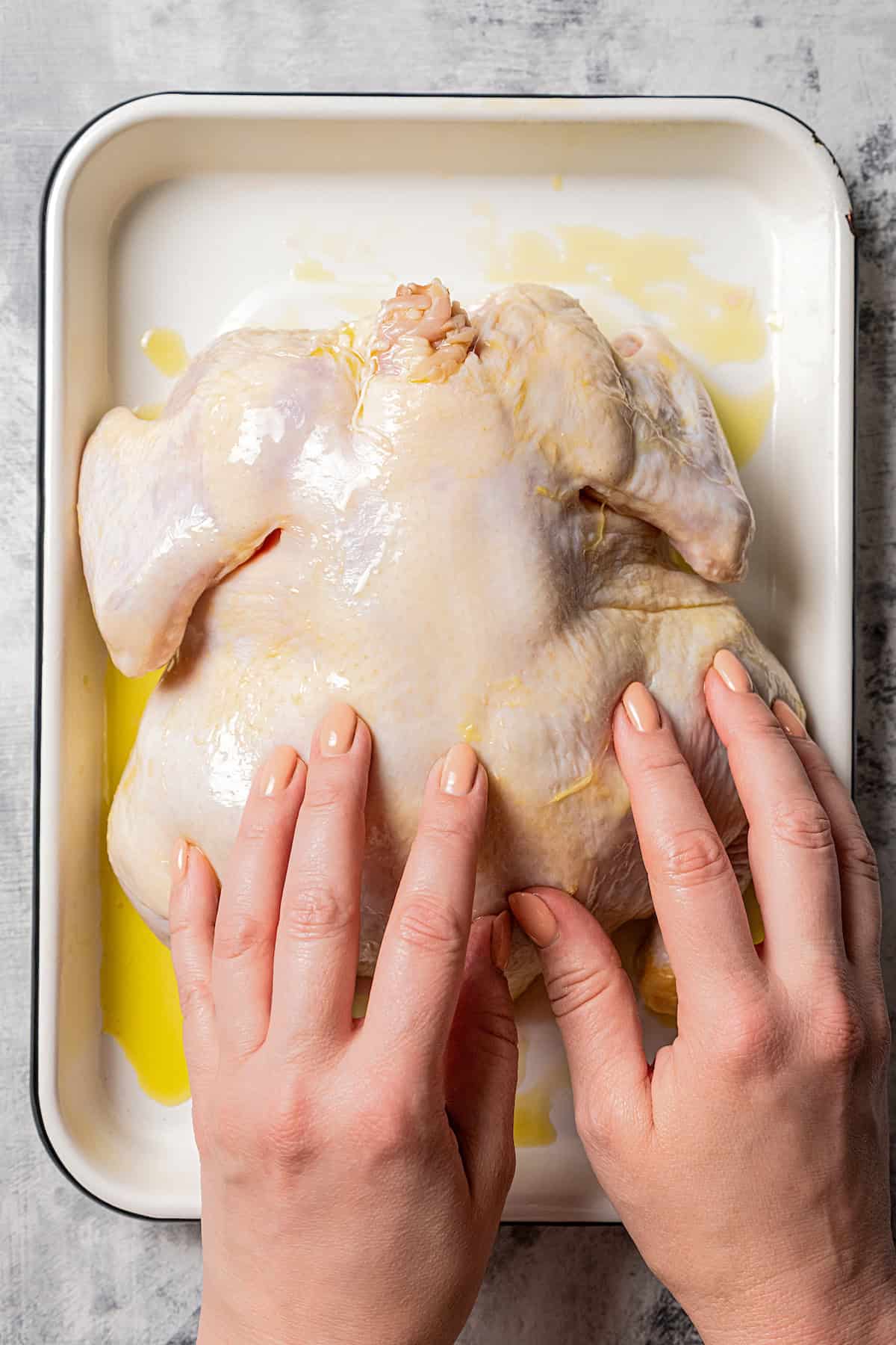 Two hands rubbing a whole chicken with oil in a baking dish.