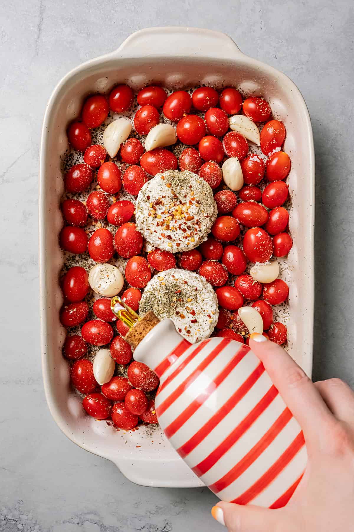 A hand holding a bottle drizzling olive oil over tomatoes, garlic, and Boursin cheese in a baking dish.