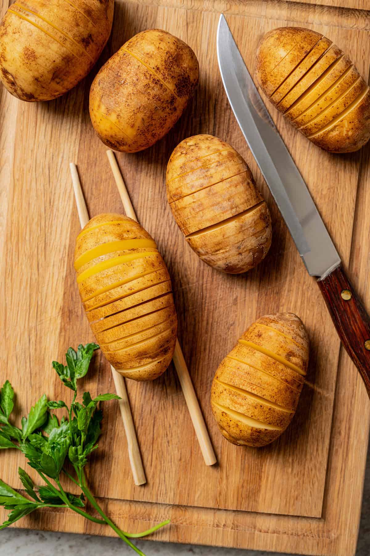 Overhead view of a sliced hasselback potato resting between two chopsticks, surrounded by more hasselback potatoes and a knife on a wooden cutting board.