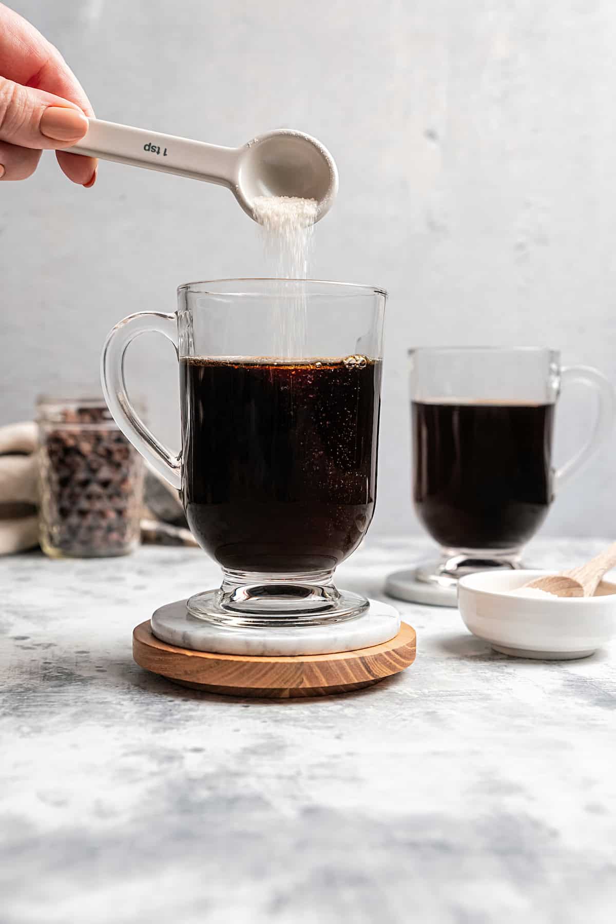 A spoonful of sugar being added to an Irish coffee in a glass mug, with a second coffee in the background.