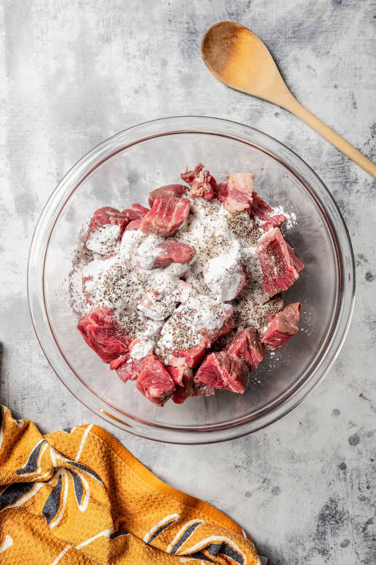 Cornstarch and seasoning added to cubed beef in a glass bowl.