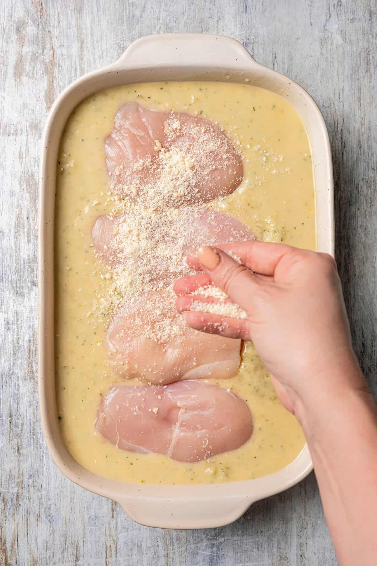 A hand sprinkling parmesan cheese over chicken nestled in a saucy rice mixture in a casserole dish.