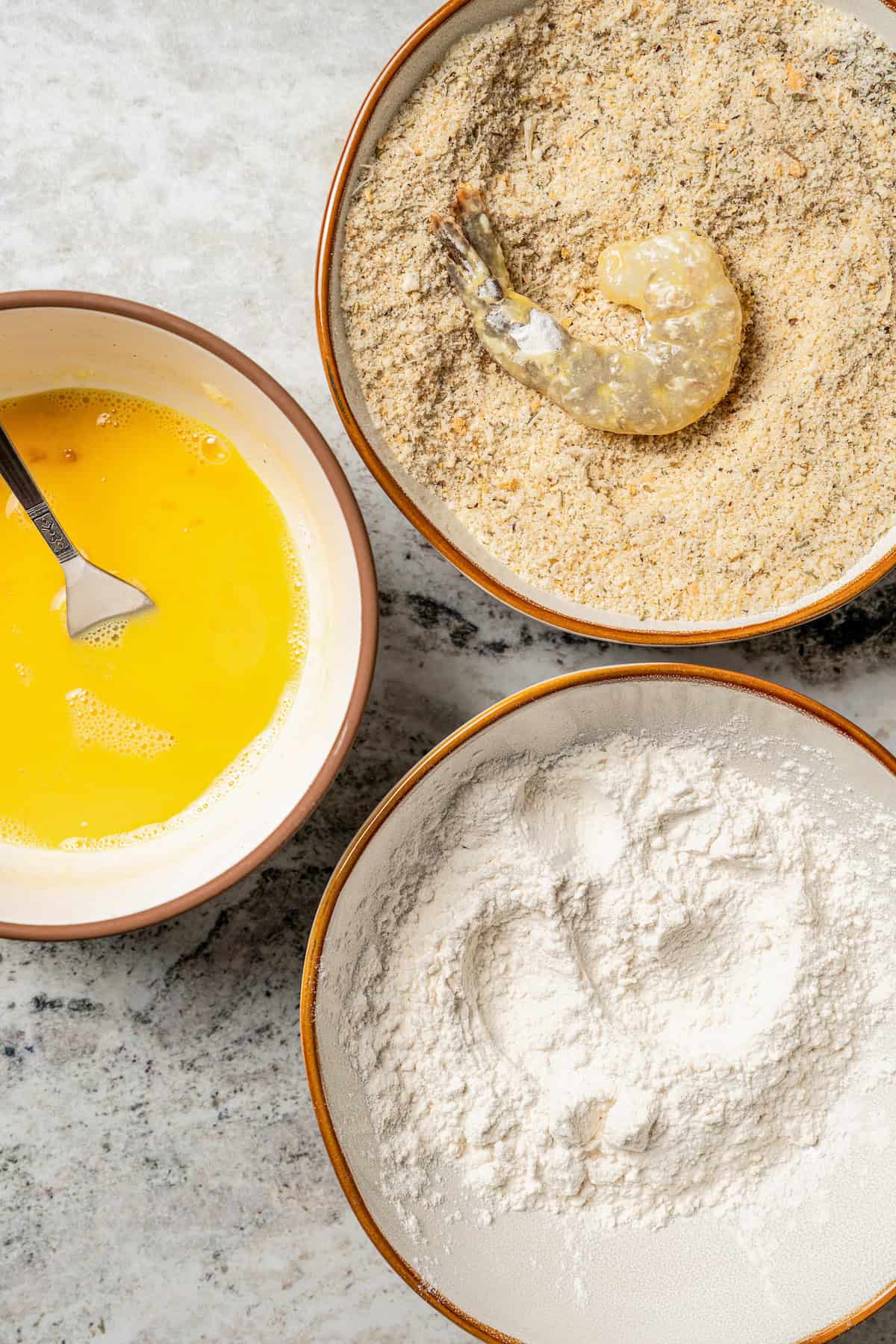 Overhead close up view of a breading station with bowls of breadcrumbs, flour, and egg.