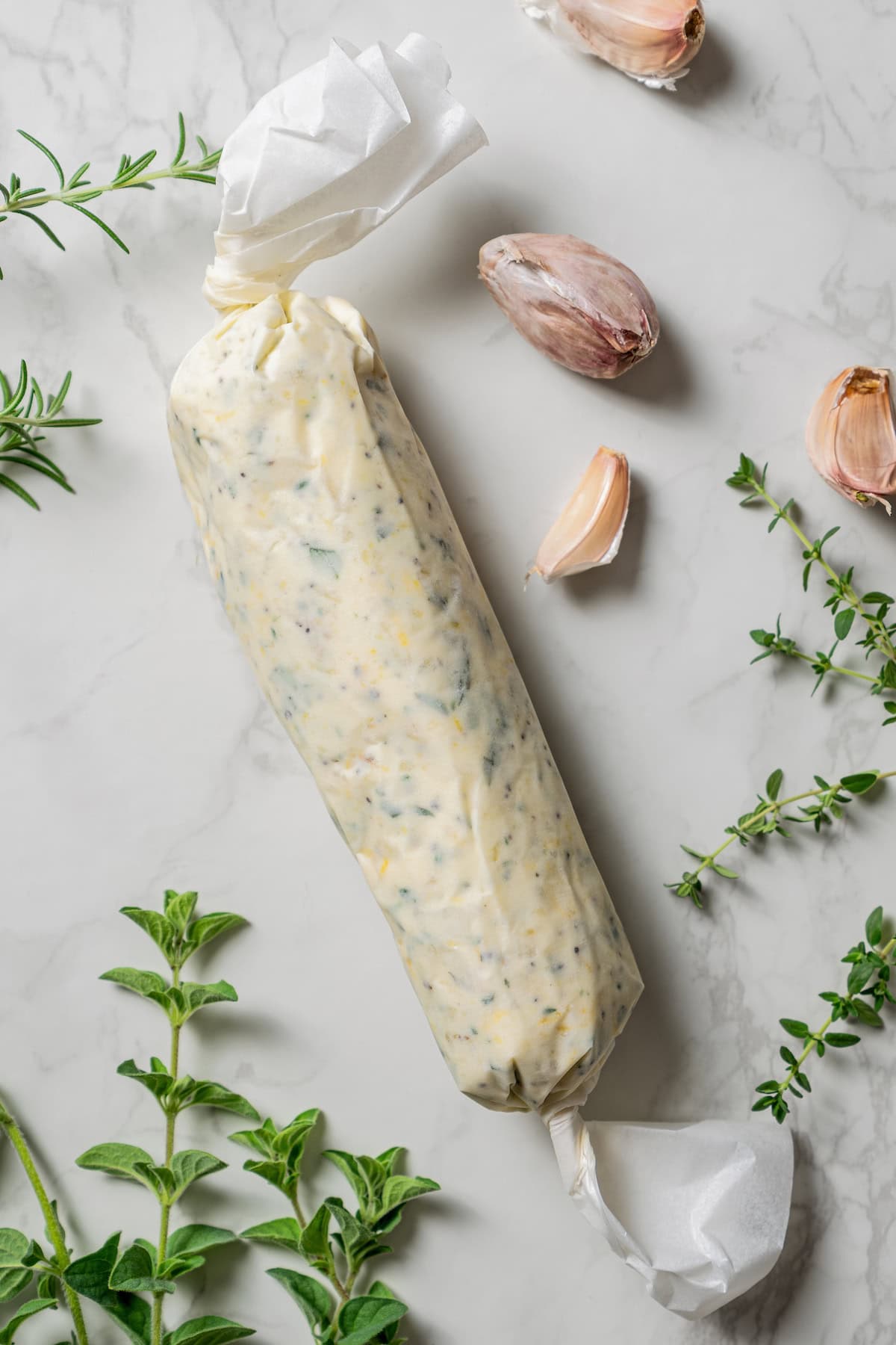 Overhead view of herb compound butter wrapped in plastic wrap to form a tight log, surrounded by fresh herbs on a countertop.