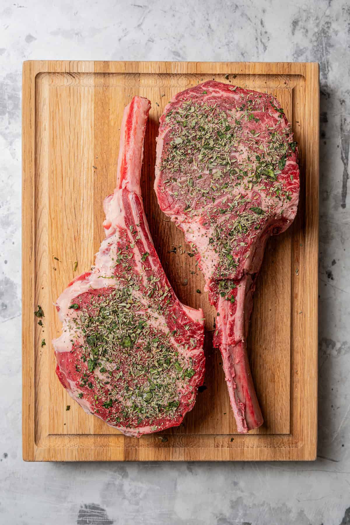 Overhead view of two seasoned uncooked tomahawk steaks on a wooden cutting board.