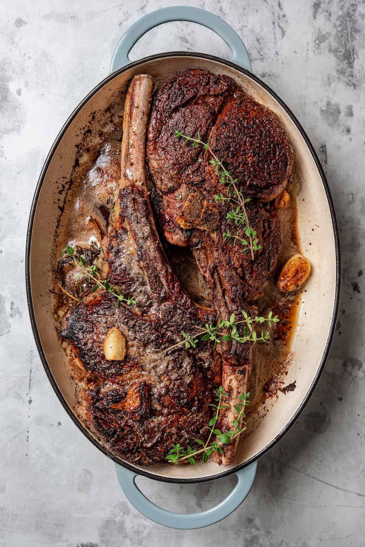 Overhead view of two seared tomahawk steaks in a large oval skillet.