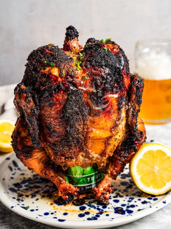 A whole-roasted beer can chicken propped upright on a plate next to one half of a lemon, with a glass of beer in the background.