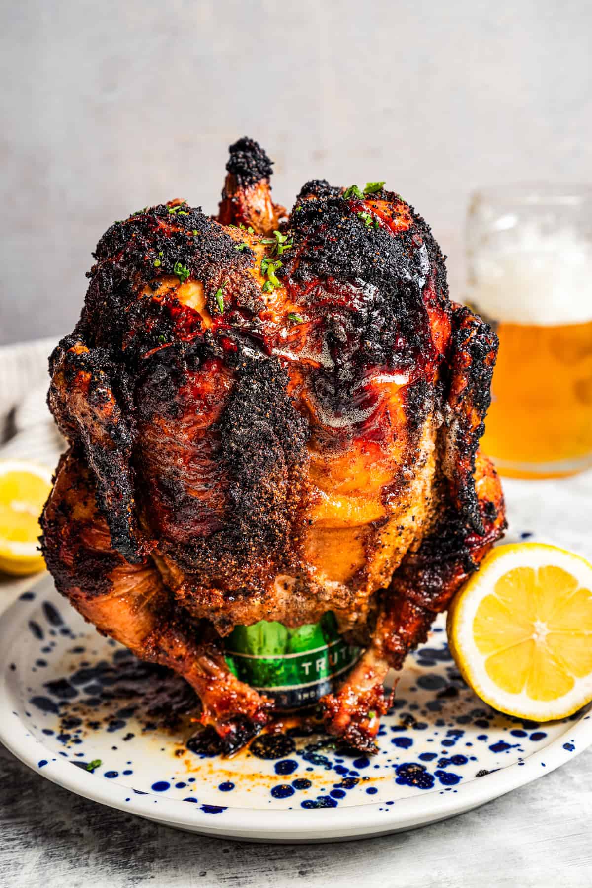 A whole-roasted beer can chicken propped upright on a plate next to one half of a lemon, with a glass of beer in the background.