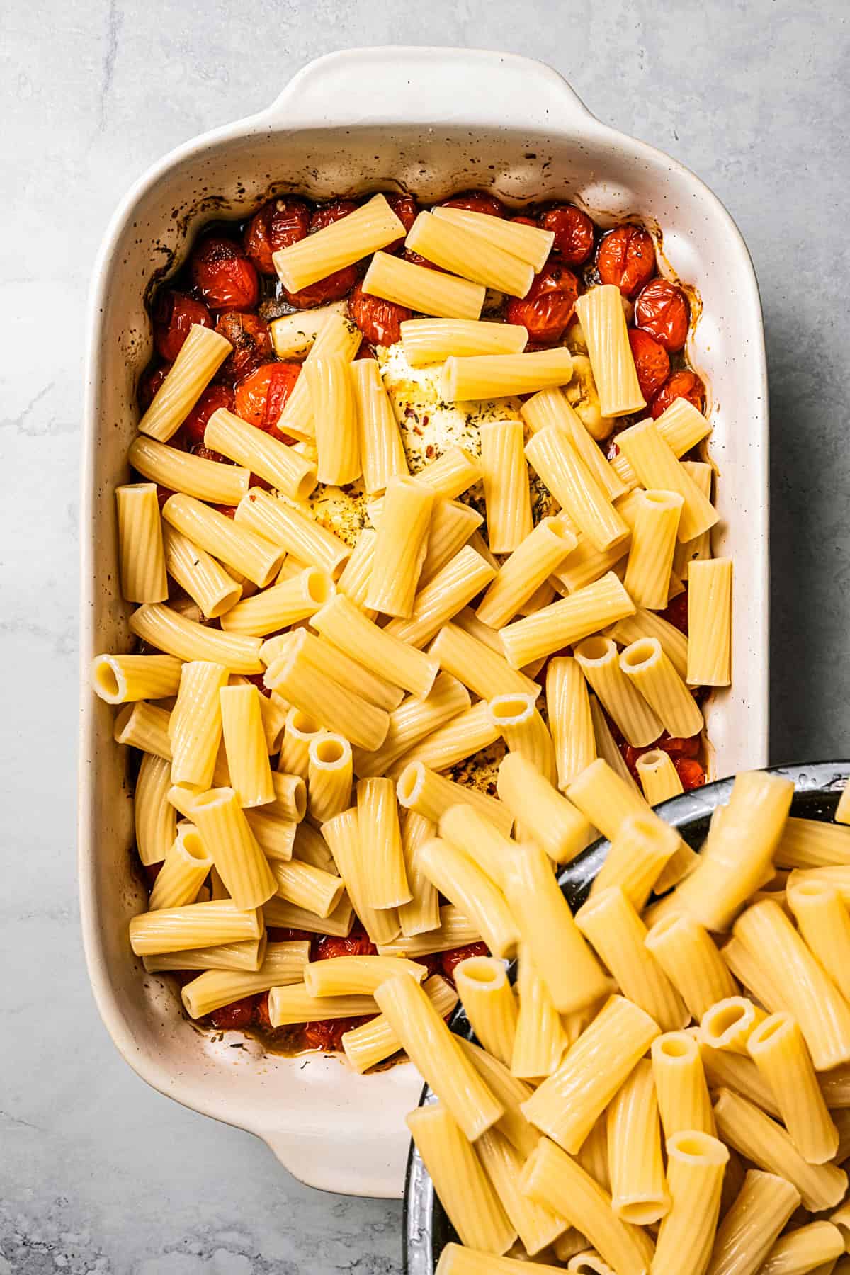 Adding boiled rigatoni pasta into a baking dish with tomatoes.