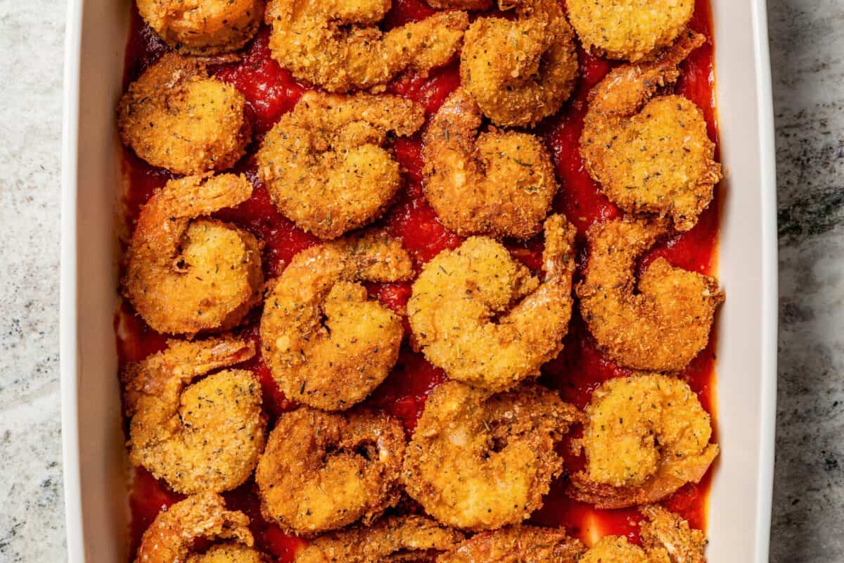 Shrimp parmigiana is crispy breaded shrimp baked over tangy marinara sauce with a golden, cheesy mozzarella crust. Perfect served with pasta! Breaded shrimp arranged over pasta sauce in a baking dish.
