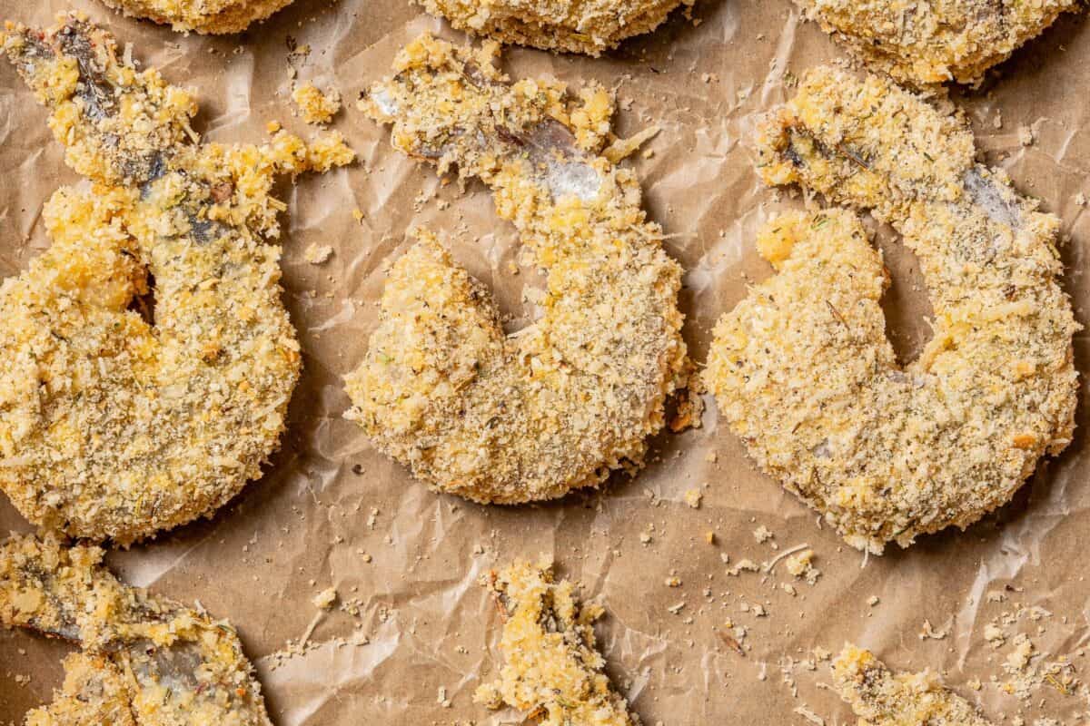 Shrimp parmigiana is crispy breaded shrimp baked over tangy marinara sauce with a golden, cheesy mozzarella crust. Perfect served with pasta! Breaded raw shrimp on a piece of parchment paper.