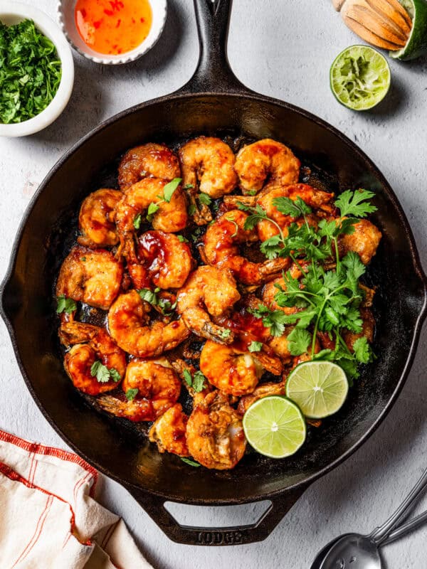 Overhead view of firecracker shrimp in a skillet garnished with cilantro and lime leaves, surrounded by small bowls of ingredients.