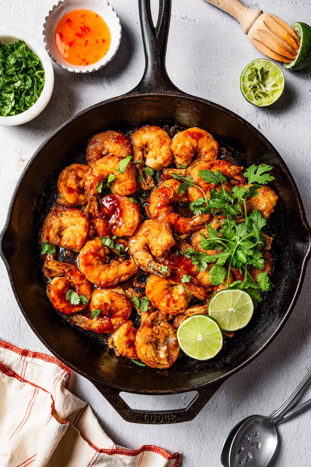 Overhead view of firecracker shrimp in a skillet garnished with cilantro and lime leaves, surrounded by small bowls of ingredients.