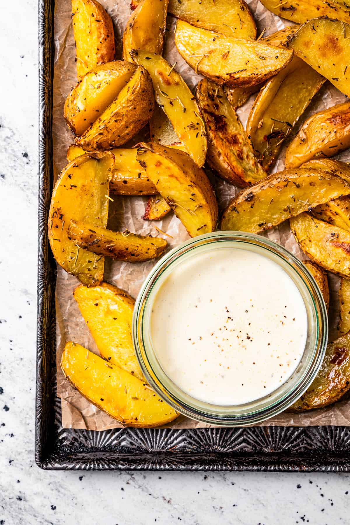 Overhead view of a small bowl of roasted garlic aioli surrounded by potato wedges on a tray.