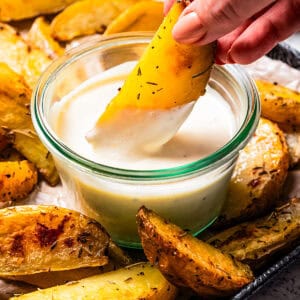 A hand dipping a potato wedge into a bowl of roasted garlic aioli, surrounded by more wedges on a tray.