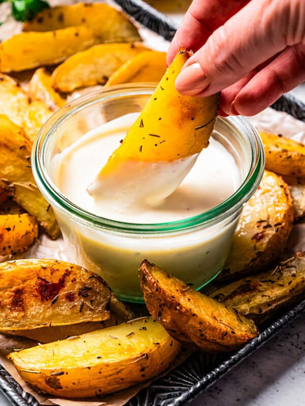 A hand dipping a potato wedge into a bowl of roasted garlic aioli, surrounded by more wedges on a tray.