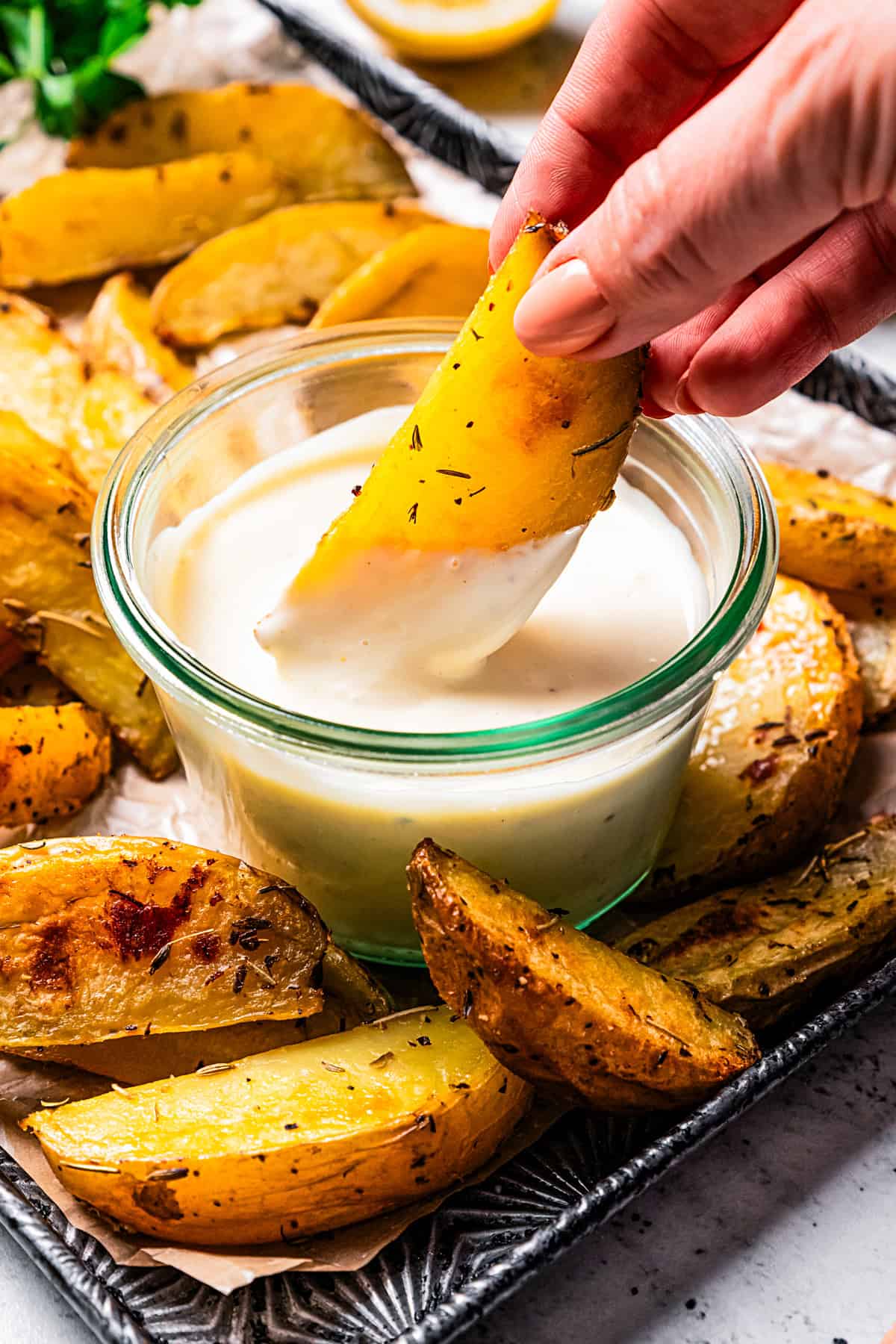 A hand dipping a potato wedge into a bowl of roasted garlic aioli, surrounded by more wedges on a tray.