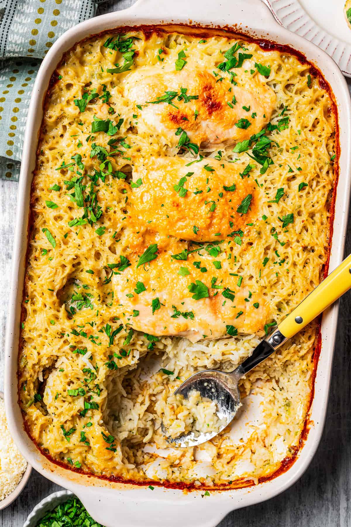 Overhead view of a baked no peek chicken and rice casserole garnished with chopped parsley, with a serving spoon resting in the space where a chicken breast was served.