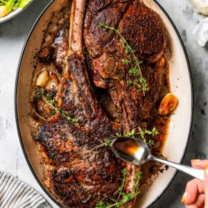 A hand spooning pan juices over two whole tomahawk steaks in a large oval skillet.