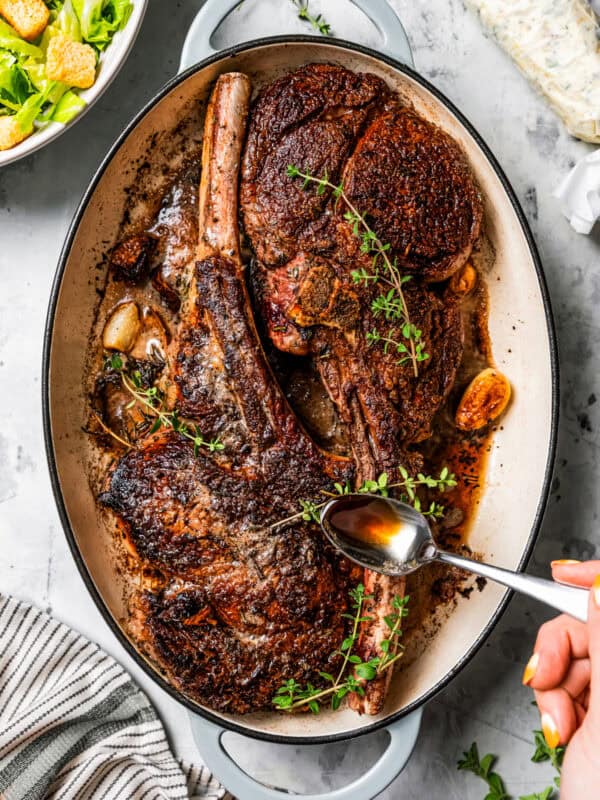 A hand spooning pan juices over two whole tomahawk steaks in a large oval skillet.