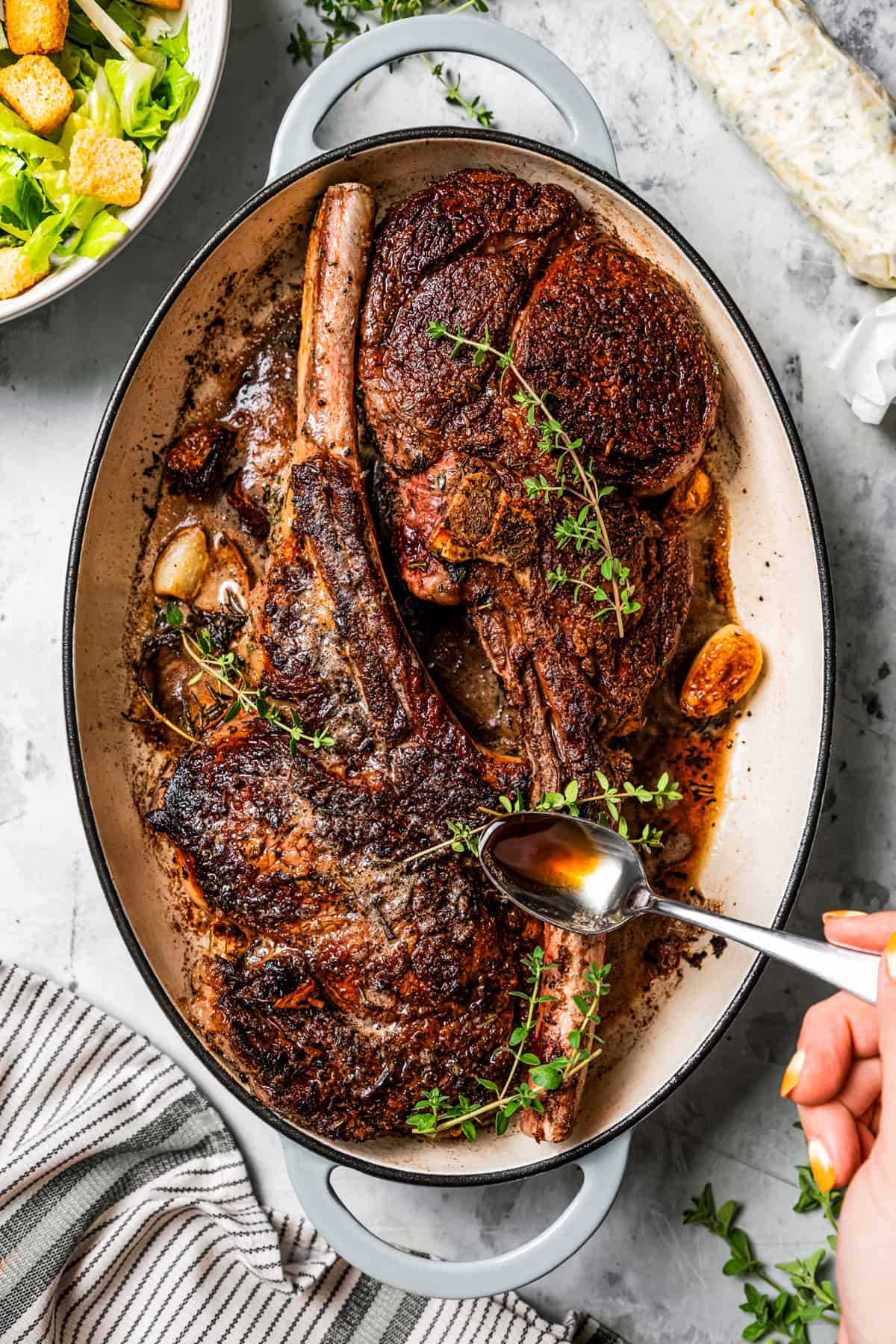 A hand spooning pan juices over two whole tomahawk steaks in a large oval skillet.